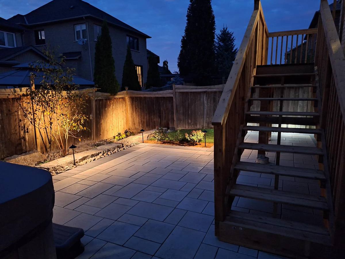 Backyard paver patio with hot tub and landscape lighting at dusk